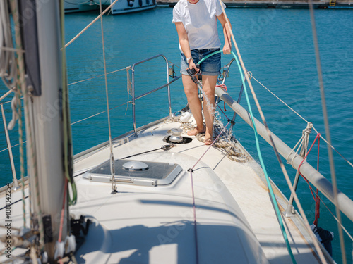 Adult woman washes teak deck of sailing yacht using water hose. Maintenance routine in Finike marina ensures boat remains clean during sunny summer day in Mediterranean Turkey.