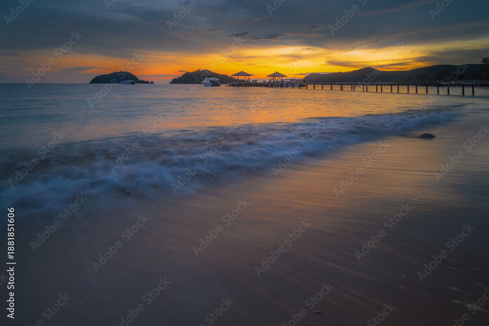 Fototapeta premium A beach with calm sea waves in the evening, a bridge in the background, and a sky with the sun setting.
