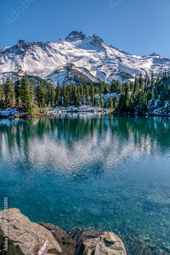 A mountain range is reflected in a lake. The water is clear and calm. The sky is blue and there are no clouds