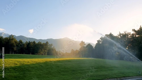 Green grass is being watered by sprinklers at a golf course during a sunny afternoon in summer.