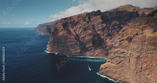 Tenerife: Los Gigantes cliffs forming the dramatic coastline, with deep blue Atlantic Ocean foregrounding towering rock faces and a small boat navigating the water. Aerial view drone panorama