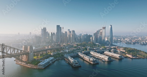 Australia, Sydney: Aerial view of Sydney Harbour Bridge and city skylines skyscrapers reflecting in water at misty sunrise, Barangaroo Reserve and wharves in foreground. Drone flight panorama
