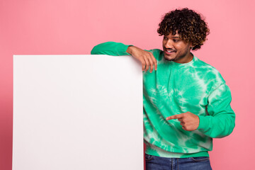 Young man with curly hair in a green tie dye sweater stands beside a blank white board smiling against a pink background