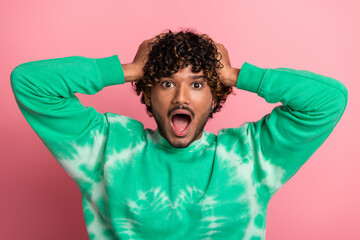 Young man in green tie dye sweater poses with hands on head against pink background