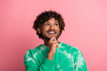 Young man with curly hair wearing a green tie dye sweater smiles and thinks against a pink background for a fashion lifestyle stock photo