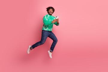 Young man jumps in the air while holding a smartphone against a pink background wearing a green tie dye sweater jeans and white sneakers