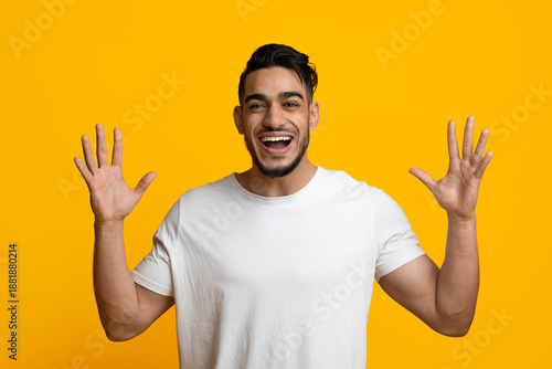 Emotional handsome millennial middle eastern man with stylish hair and beard raising hands up and smiling, expressing amazement on yellow studio background, human gestures and emotions concept