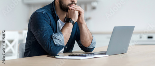 Upset, sad, depression, frustrated and work at home. Concentrated pensive millennial bearded male looks at laptop and pressed hands to mouth, sits at table in kitchen interior, profile, panorama