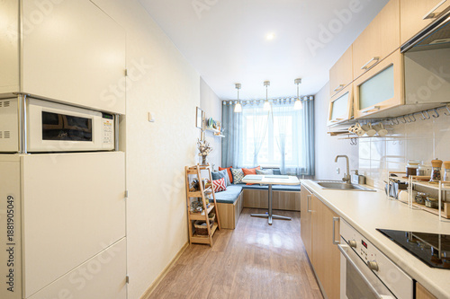 narrow, modern kitchen with light wood cabinets, white appliances, sink, microwave, dining nook with cushions and pillows, pendant lights, and a shelving unit. Bright and cozy.