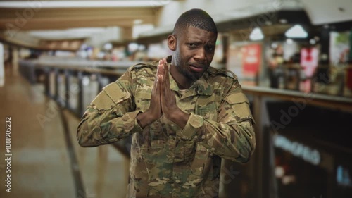 Soldier man in camouflage uniform pressing hands together near storefronts on airport concourse in pleading gesture; plea anxiety.