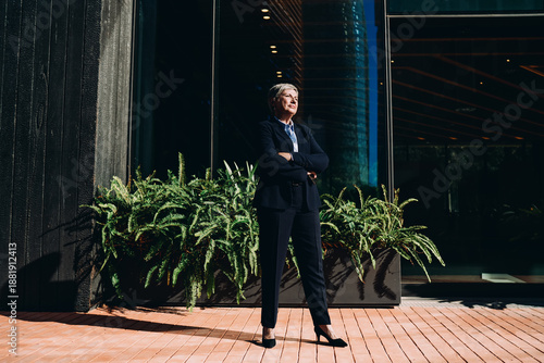 Quadro em tela Senior businesswoman standing proudly outside modern building with arms crossed, showcasing strategic confidence, independence, and executive presence in digital context