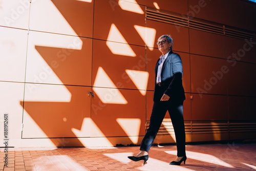 Принти на полотні Senior professional woman striding purposefully under dramatic shadows, symbolizing ambition, resilience, and forward motion in a tech-connected urban world