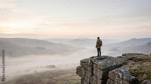 A lone backpacker stands on a rocky cliff, gazing out over a misty valley at dawn. The low clouds create an ethereal, atmospheric scene in the Peak District National Park.