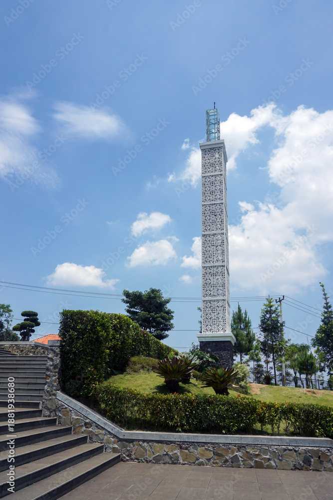 Fototapeta premium The tall mosque tower with modern white architecture stands firmly in the neatly maintained mosque garden area, with a backdrop of blue skies and bright clouds. 