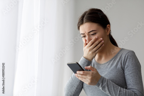 Fotografi Upset depressed millennial pretty woman covering her mouth with hand and looking at phone, got bad news at home