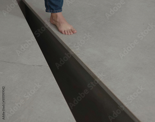 A bare foot wearing denim approaches the sharp edge of a linear, dark void separating gray tiled flooring.