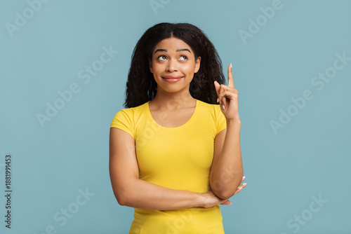 Fotografie Black lady pointing finger up and looking upwards, having idea, found solution or got inspiration, standing on blue background, studio shot