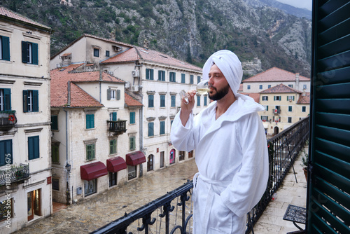 Man in white bathrobe and towel on his head stands on balcony overlooking historic European old town and drinks from wine glass. Buildings with shuttered windows and red roofs at background