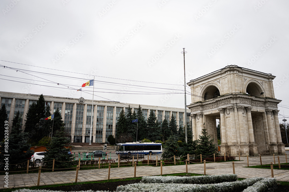 Obraz premium Ornate Triumphal Arch monument beside a government building in Chisinau Moldova with trees and people