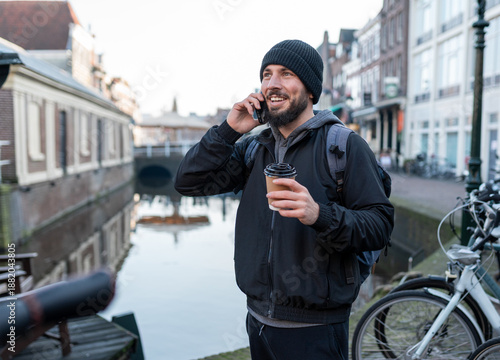 Young man using smartphone and drinking coffee by delft canal, enjoying urban communication and travel