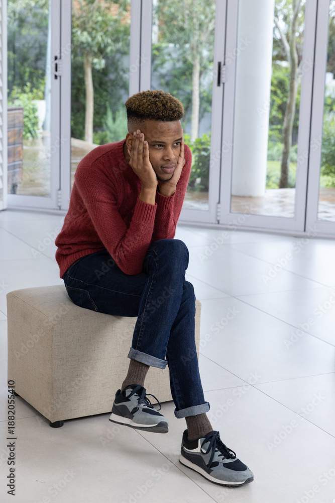 Naklejka premium African American male sitting on ottoman in bright room in red sweater, gazing through glass doors