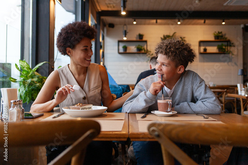 African American woman and her son are sitting in cafe drinking coffee and smiling, curly-haired teenager and his mother are having lunch in restaurant