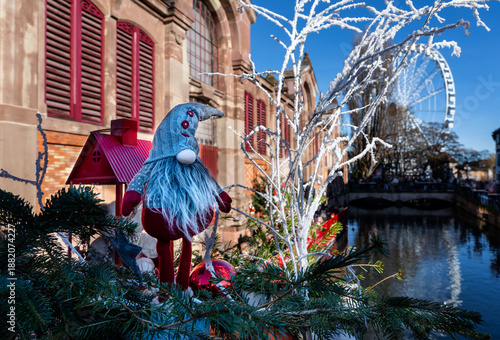 In the foreground, Christmas decorations with a cute gnome and white branches; in the background, a canal in Colmar, France, under a blue sky.