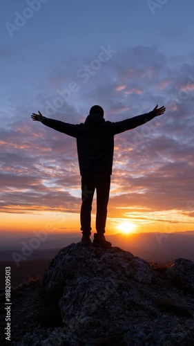 A person stands on a rock with arms outstretched at sunset.