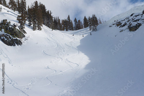 Striking alpine scene in a snowy mountain bowl featuring winding ski tracks descending the right side of the slope, contrasted by clusters of roller balls (snow rollers) forming on the left under sun.