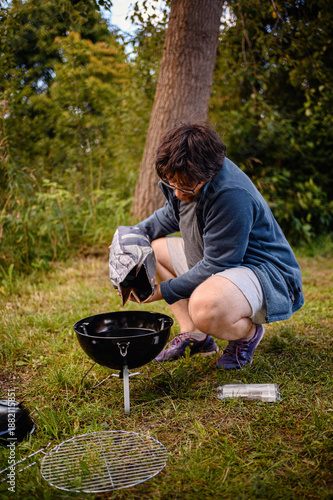Adult camper crouches on grass and pours charcoal into a small portable kettle grill at a wooded campsite, capturing outdoor cooking, camping gear, and weekend travel vibes.