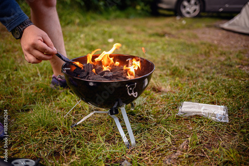 Close-up of a camper using tongs to stoke burning charcoal in a small kettle grill on grass, with campsite gear in the background—ideal for outdoor cooking and travel themes.
