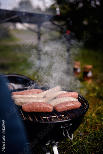 Various sausages cook over a smoky charcoal grill outdoors, with blurred jars and greenery in the background.
