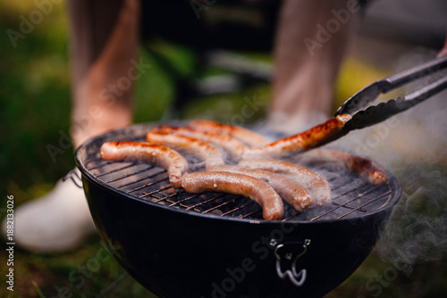 A close-up of tongs flipping a grilled sausage over hot charcoal with visible smoke rising during a campsite barbecue.