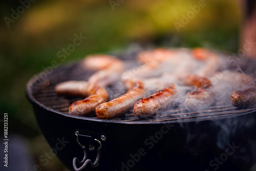 Grilled sausages sizzle with visible smoke rising from a hot charcoal grill during an outdoor cookout.