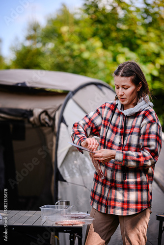 A woman in a plaid shirt opens packaged meat while preparing a meal at a camping setup outdoors.
