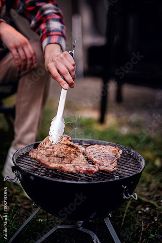 A close-up of a person grilling seasoned steaks on a portable charcoal grill during a camping trip.