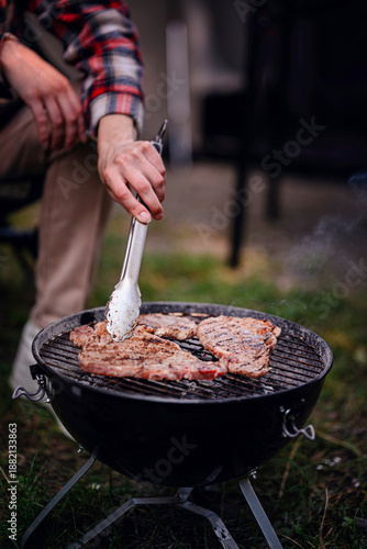 A close-up of a person grilling seasoned steaks on a portable charcoal grill during a camping trip.