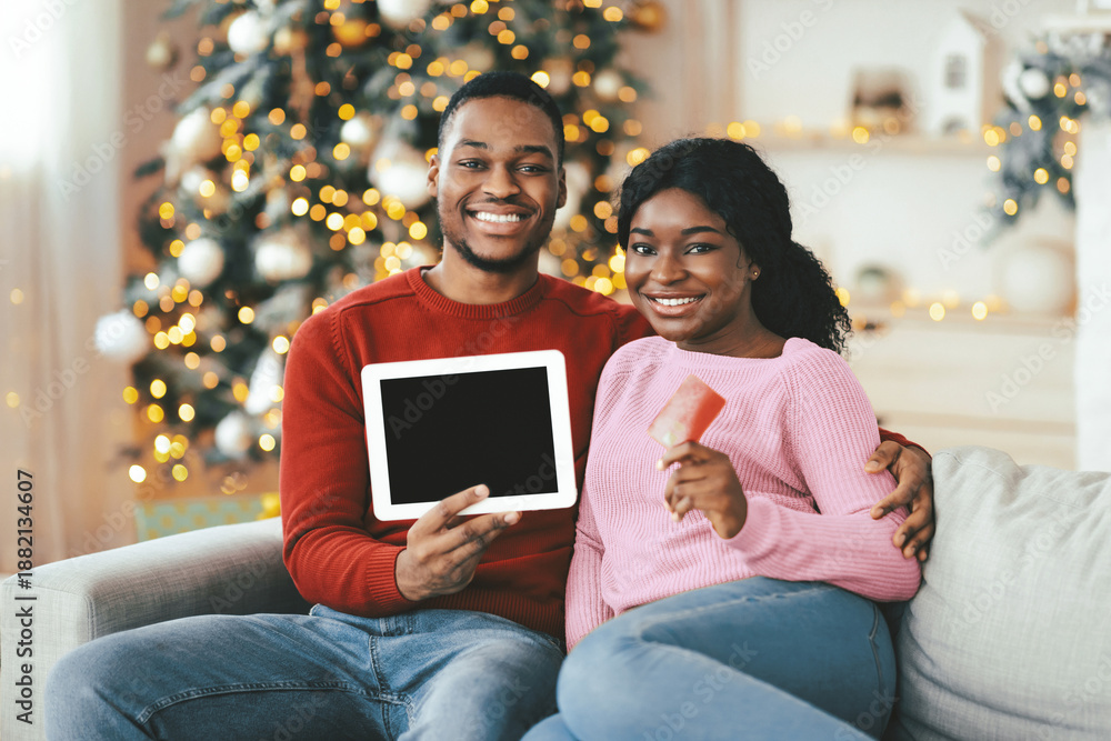 Obraz premium Couple sits on a couch holding a tablet and a gingerbread cookie. They enjoy holiday decorations in their cozy living room. A Christmas tree shines in the background.