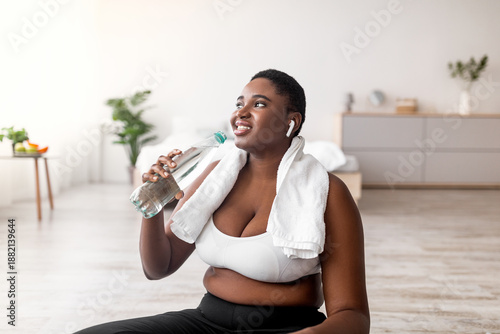 Photos Portrait of young plus size black woman with earphones drinking water and listening to music on break from home training