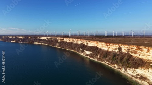 Wallpaper Mural Cliffs rise above the water with green vegetation along the shore. Wind turbines stand tall in the background against a clear blue sky on a sunny day. Torontodigital.ca