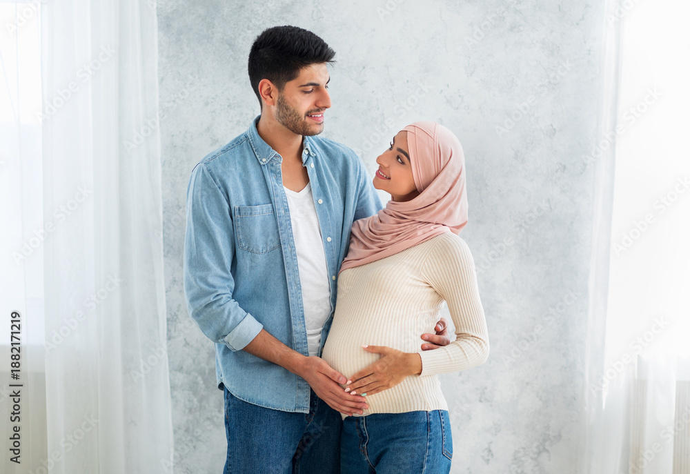 Fototapeta premium Happy parents-to-be. Happy muslim couple waiting for baby, man embracing wife and touching tummy, standing at home interior, free space. Islamic spouses cuddling and enjoying future parenting