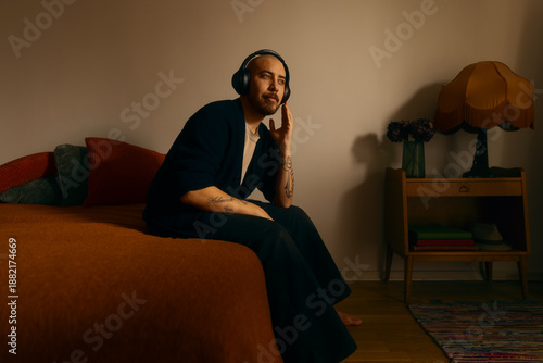 Quadro em tela A thoughtful man wearing wireless headphones sits on a bed in a warm, vintage inspired room