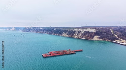 A mussel farm sits in the water with a structure for growing shellfish. The scene includes a hilly shore and wind turbines in the distance under cloudy skies.