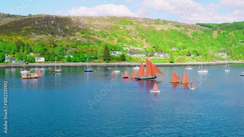 Aerial DJI Inspire drone view of Ullapool, Scotland, during summer festival, showing traditional lugger boats on calm harbor waters, festival crowds, colorful tents, scenic Scottish coastline, bright 