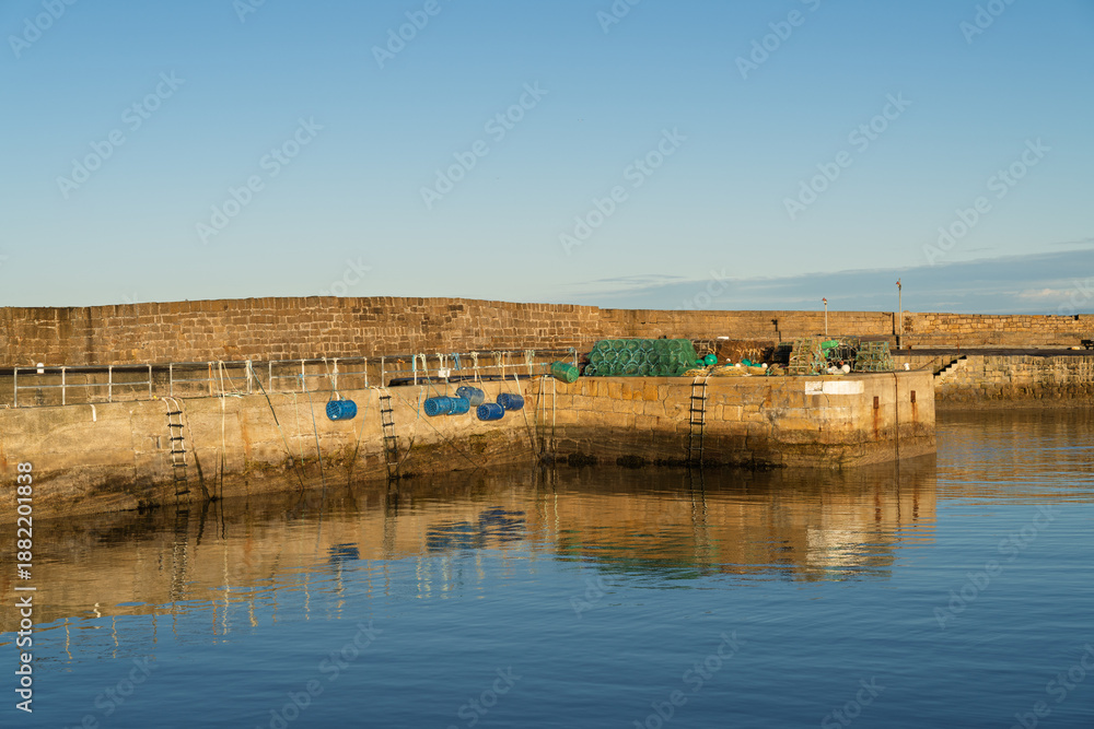 Fototapeta premium Golden hour sunlight shines on quay at Hopeman Harbour revealing still water reflections
