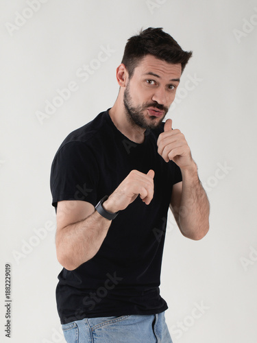 Tableau sur toile Expressive young man in black t-shirt standing in a defensive boxing pose, looking at camera with funny defiant face, grey background