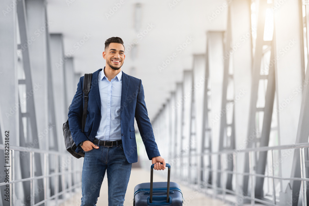 Fototapeta premium Portrait Of Handsome Happy Arab Man Walking In Airport Terminal With Suitcase, Smiling Young Middle Eastern Guy In Casual Clothes Going To Boarding Gate, Enjoying Upcoming Air Trip, Copy Space