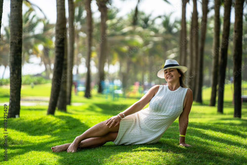 Fototapeta premium A girl in a white dress and hat lies on a green lawn against a background of palm