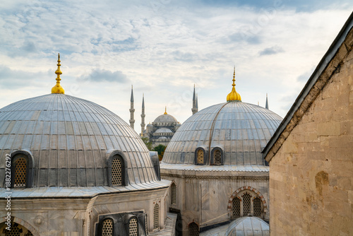 Historic mosque domes with golden finials and minarets in the background under a cloudy sky, representing Ottoman architecture, Islamic culture, and Istanbul cityscape.