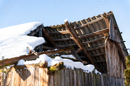 Damaged wooden barn roof partially covered with snow against a clear blue sky, emphasizing the contrast between winter elements and structural decay. Ideal for themes of abandonment or rural life.